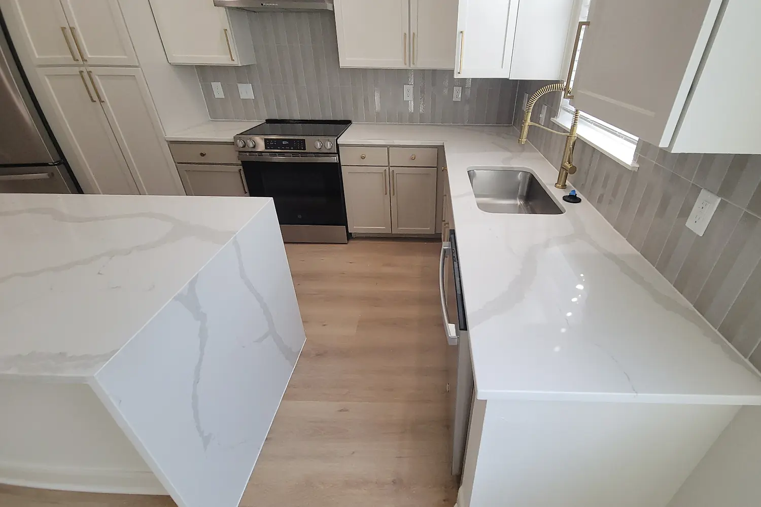 Finished kitchen with white veined countertops, waterfall island, white cabinetry, gray tile backsplash, and brass fixtures.