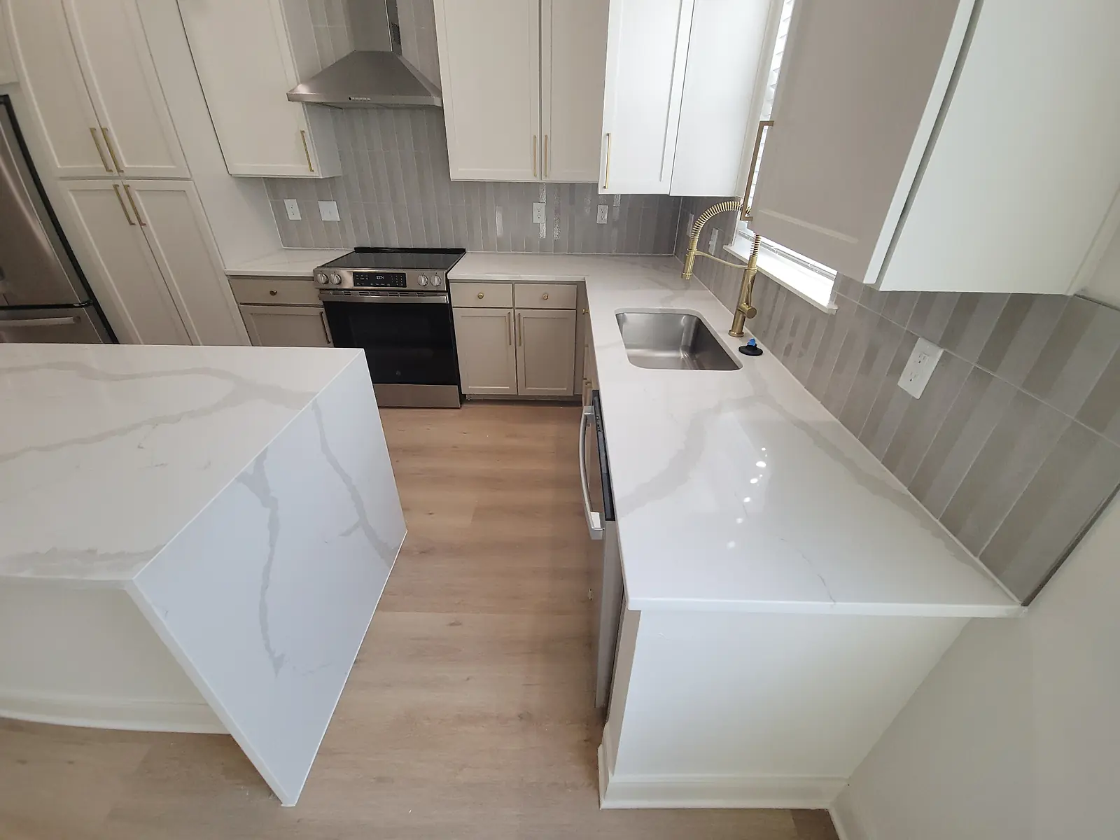 Finished kitchen with white veined countertops, waterfall island, white cabinetry, gray tile backsplash, and brass fixtures.